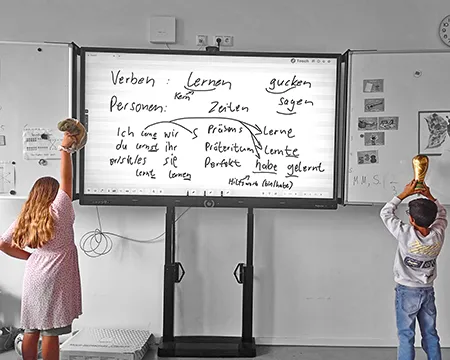 Kinder stehen mit dem Rücken zum Fotografierenden und halten vor dem Tafelbild mit deutscher Grammatik etwas hoch.